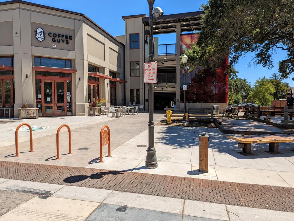 Photo of parking lot entrance behind a pedestrian plaza and attached to a coffee shop with bicycle parking in the foreground