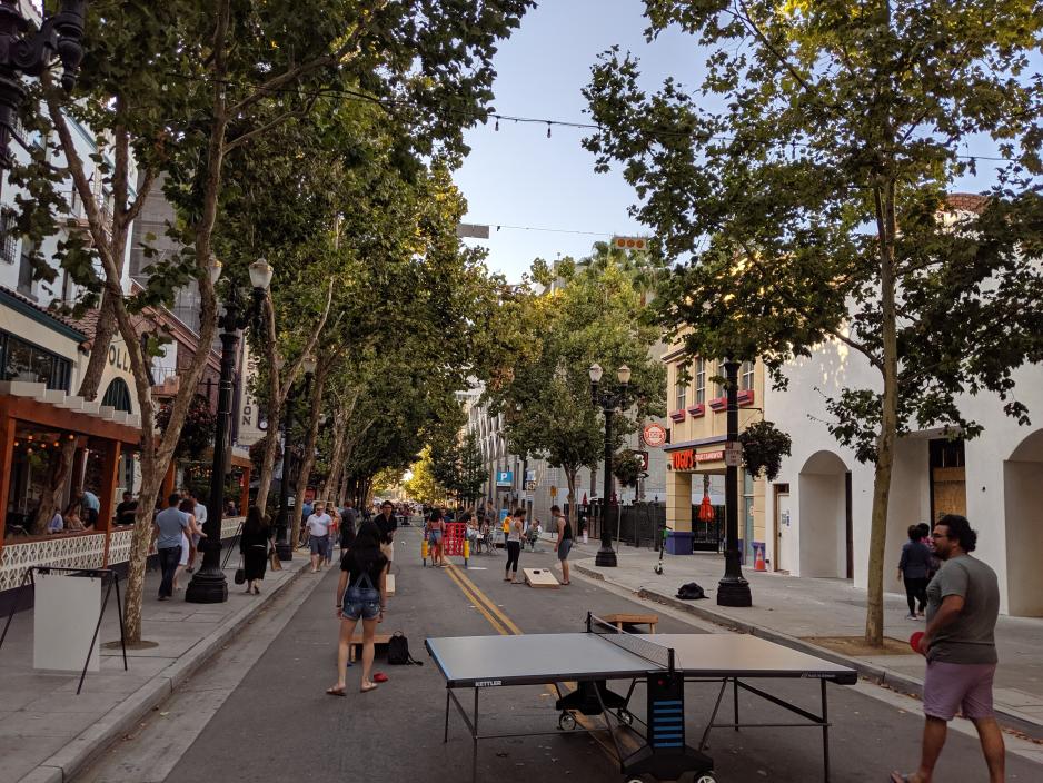 Photo of a narrow city street closed to traffic with people playing games at a ping pong table and a giant connect four game. Street trees, restaurant seating on the sidewalk and street.