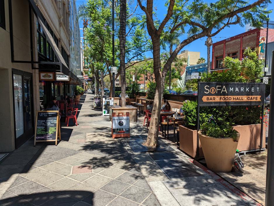 Photo of a sidewalk with the parking lane converted into a parklet with seating for adjacent businesses. Street trees.