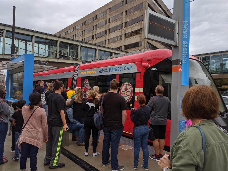 A photo of a street car with people waiting to get onboard. The stop has large, blue signs directing pedestrians to the stop. 