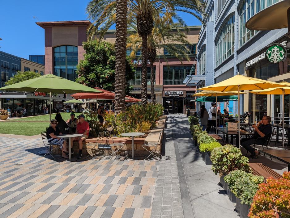 A photo of a public space surrounded by restaurants on two sides. The public space includes outdoor seating, planter boxes, and a grass lawn.