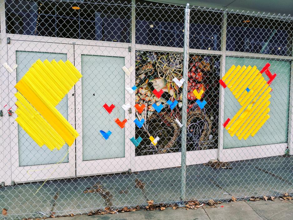 A photo of a chain link fence with a pac man and hearts art made out of tape.
