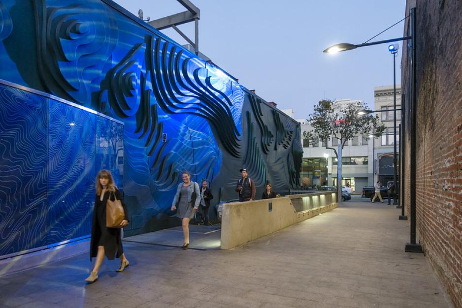 Photo of alley with blue 3-D mural on a building wall. Pedestrians walking in the alley. Evening, lights are on.