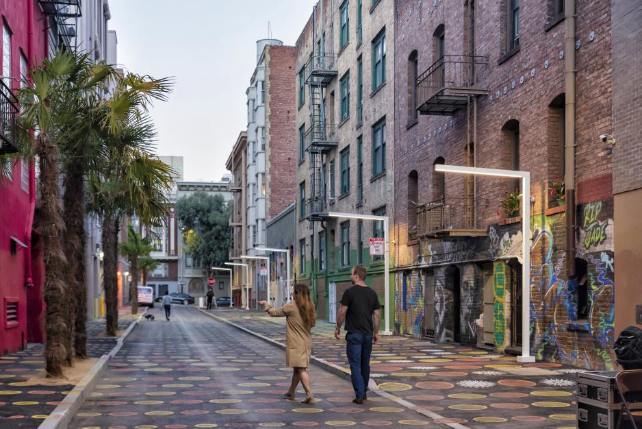 Photo of an alley with modern street lights, palm trees, polka dots on the street and walls. Two pedestrians are walking. It is daytime.