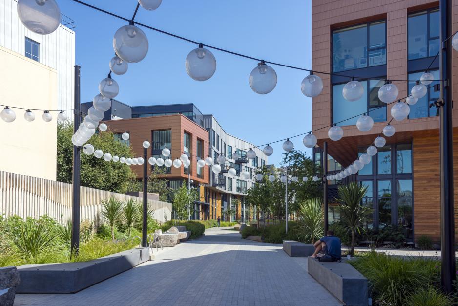 Daytime photo of alley bordered by apartments, benches, street trees. String lights cross the alley and a pedestrian is walking.