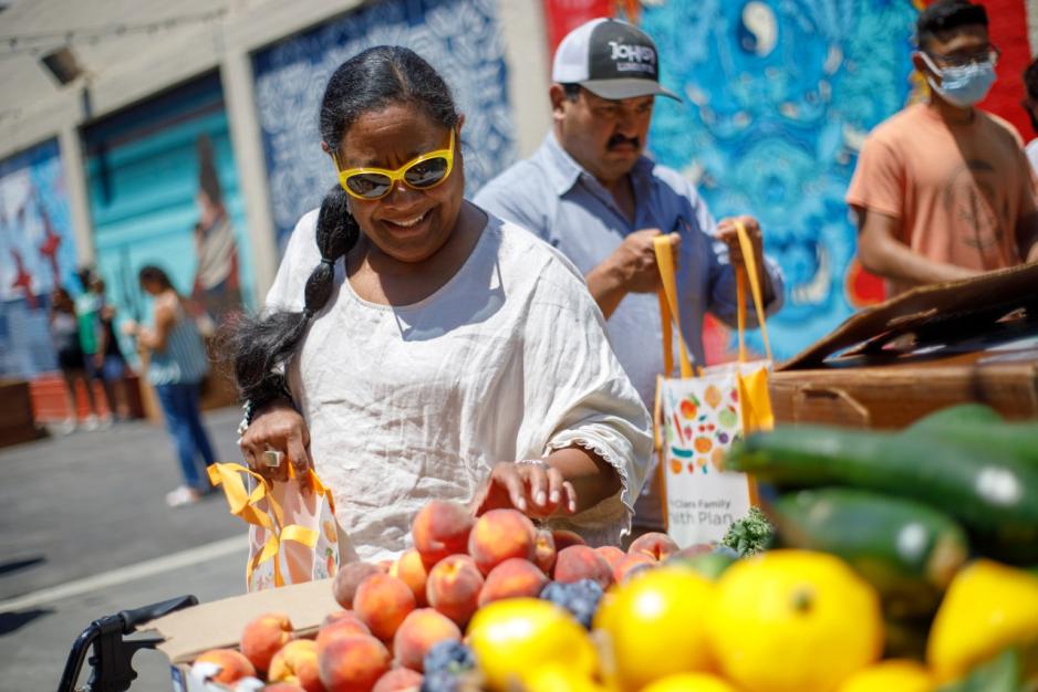 Photo of a woman smiling, reaching for an orange at a farmer's market fruit stand located on a street closed to traffic.