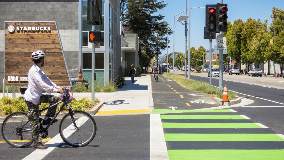 Photo of a raised bikeway on a major street, with green crossing markings, bicyclist waiting to cross the street.