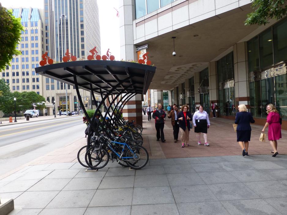 A photo of a busy and wide sidewalk adjacent to a high-rise building with shaded bike parking. 