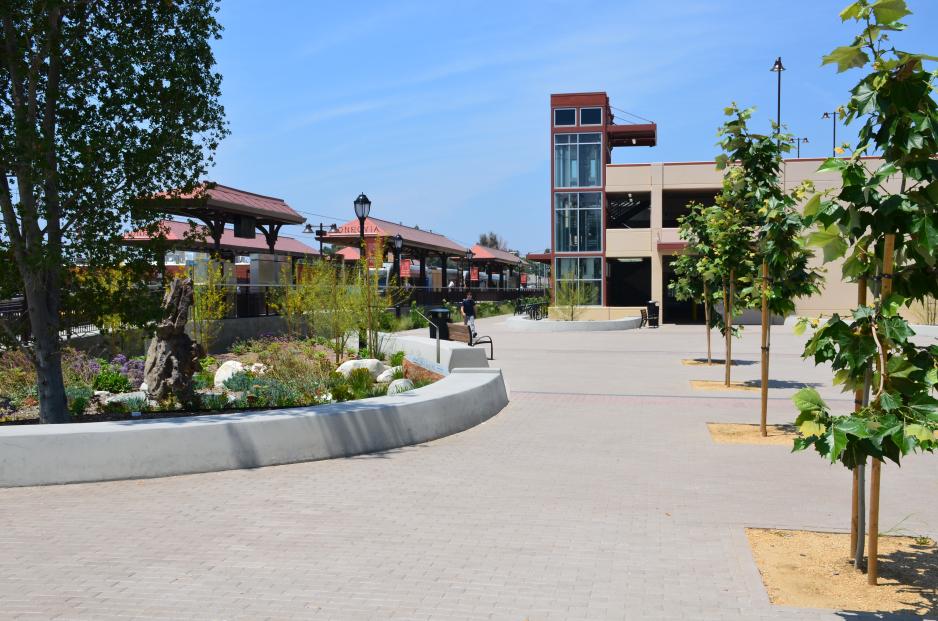 A photo of open plaza next to a transit center and parking garage. The plaza has flexible seating and trees/plants. 