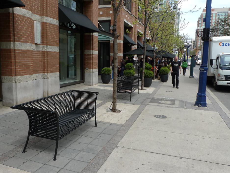 A photo of a benches on a sidewalk near trees and a cafe's patio seating.