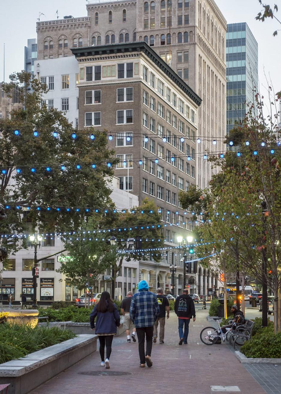 A photo of a brick sidewalk with planter box seating, trees, bike parking and lights.