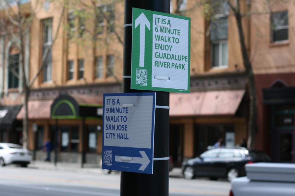 A photo of two signs on a light pole pointing pedestrians to Guadalupe River Park (a 6 minute walk) and City Hall ( a 9 minute walk).