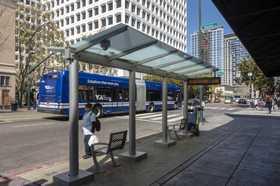 A photo of a bus shelter with people waiting, seating, trash can, and real time schedule.