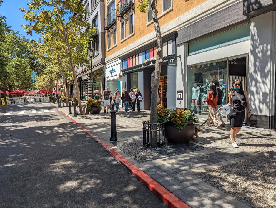 Photo of sidewalk in front of colorful shops with trees and landscaping and people walking