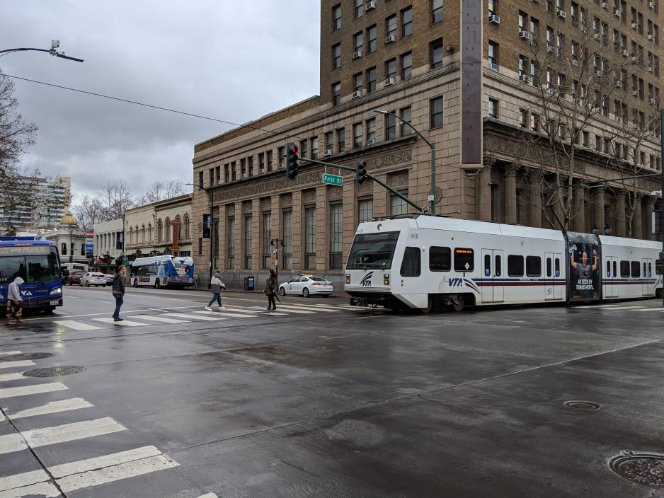 A photo of a light rail train traveling downtown near a VTA bus and people crossing the street.