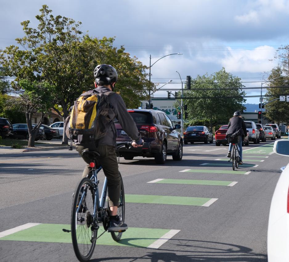 Photo of two bicyclists using a bicycle lane between a vehicle lane and parked cars on a partly sunny day