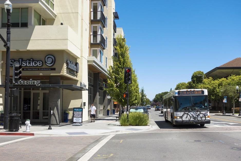 Photo of a roadway with a bus, sidewalk with landscaping, and a taller apartment building 