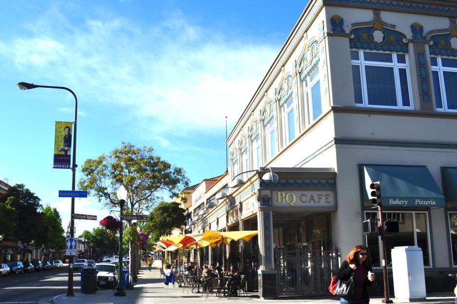 Photo of two-story building with café tables with umbrellas in front and a person walking