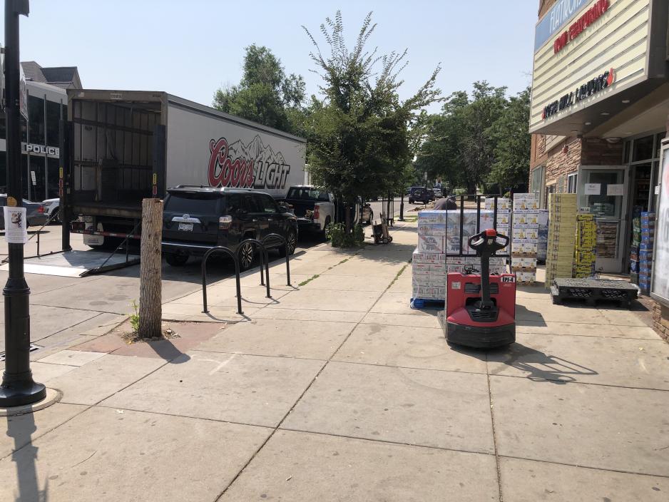 Photo of the sidewalk in front of a shop with parked cars at the curb and a delivery truck blocking them in as items are being unloaded