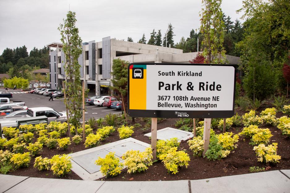 Photo of sign for park and ride area in front of the parking lot and garage with cars parked