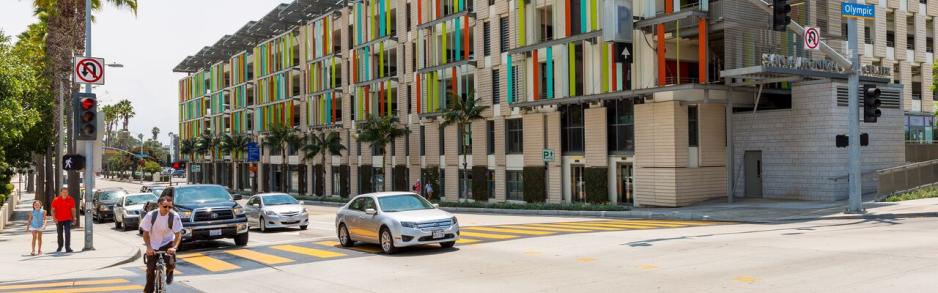 Photo of taller parking garage decorated with several colors and shopping underneath and drivers and bicyclists traveling in front