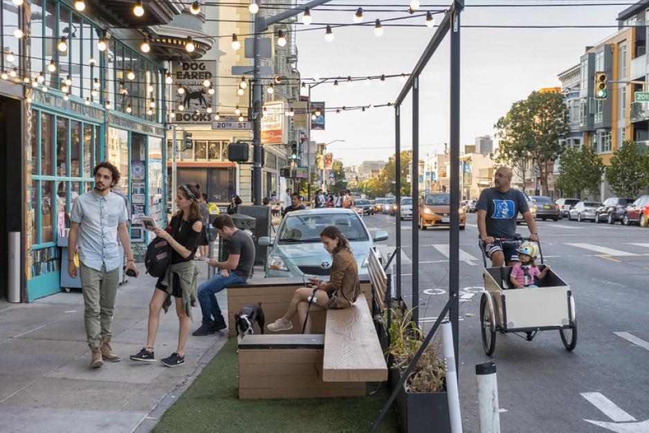 Photo of people sitting in a parklet with string lights above, fake grass, a bench, a dog, and a person biking by with a child