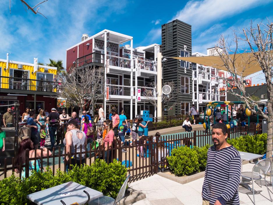 Photo of people standing and walking in front of one- to three-story buildings made from shipping containers
