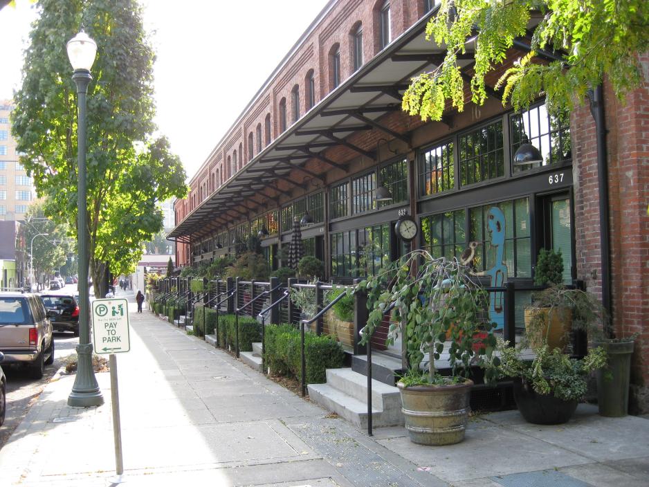 Photo of several shop entrances with trees and landscaping with parked vehicles and a sign that says "Pay to Park"