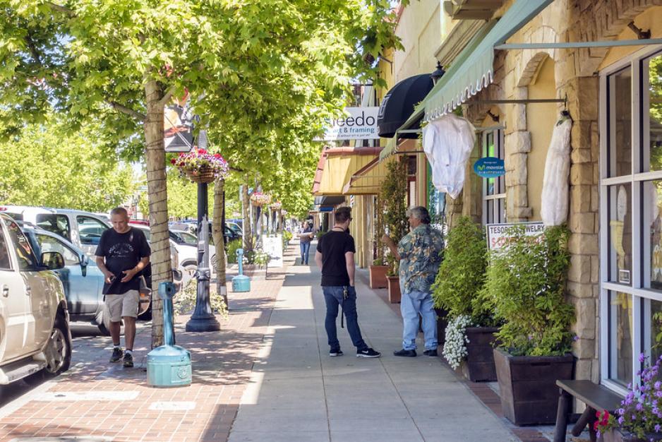 Photo of sidewalk with people standing and walking with trees between parked vehicles and store entrances open