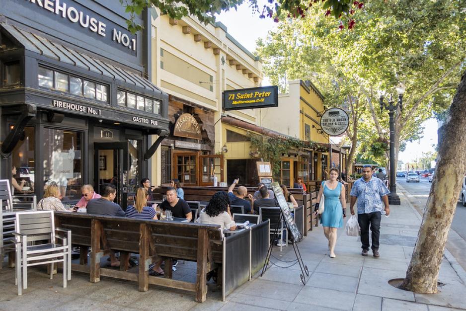 Photo of people sitting outside restaurants under trees with people walking by