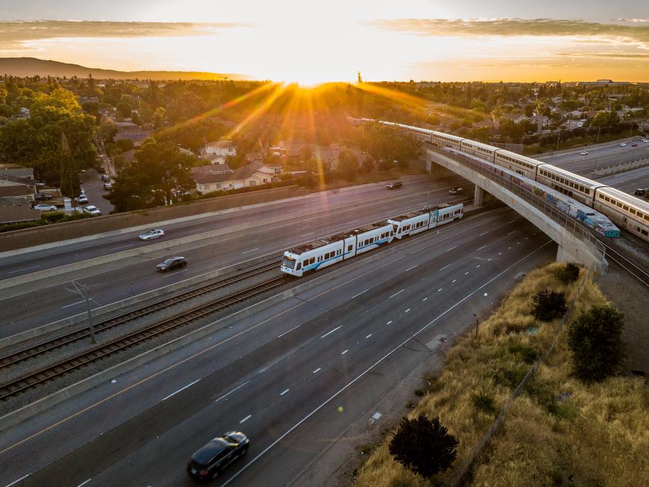 Photograph of a sunset showing light rail, a freeway, and another train passing above 