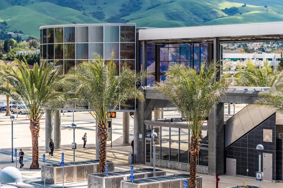 Photo of Warm Springs BART station area entrance with trees and bike lockers in the foreground with green hills and buildings in the background