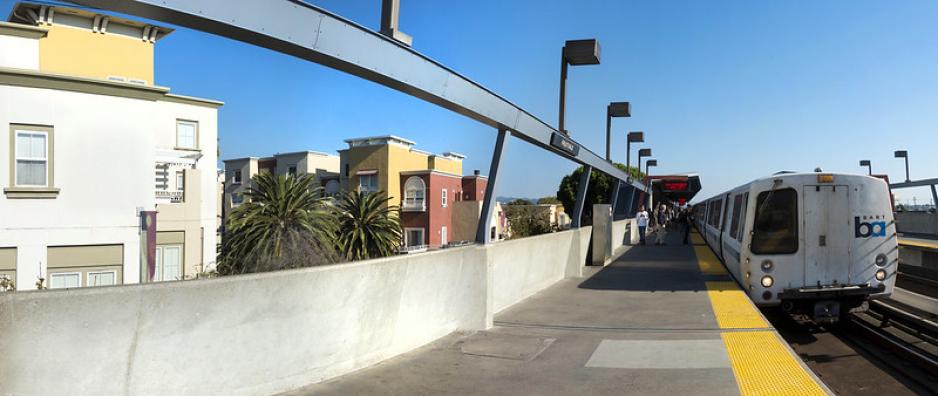 A photo of BART at an elevated station next to apartment buildings.