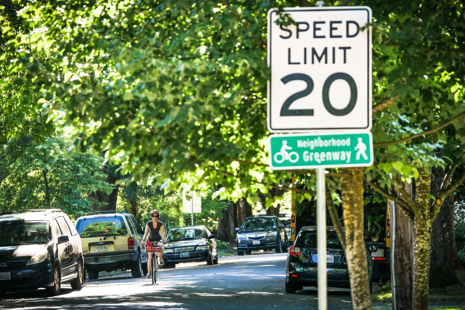 A photo of a local street with a 20 mph sign, neighborhood greenway sign, trees, and a cyclist.