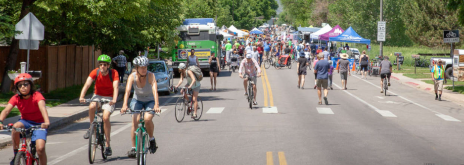 Photo of a roadway with hundreds of people walking or riding bicycles while wearing helmets with a food truck and tents in the background