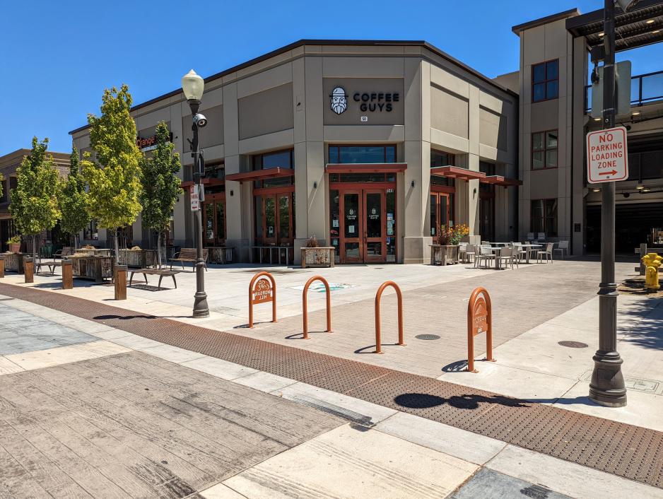 Photo of a pedestrian plaza with a tan coffee shop with multiple entrances. There are several bike racks and trees