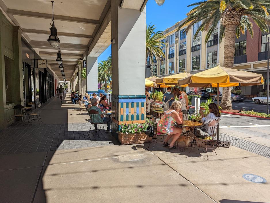 Photo of a walkway under a building with shops on the side and people sitting at tables on the other side on a sunny day