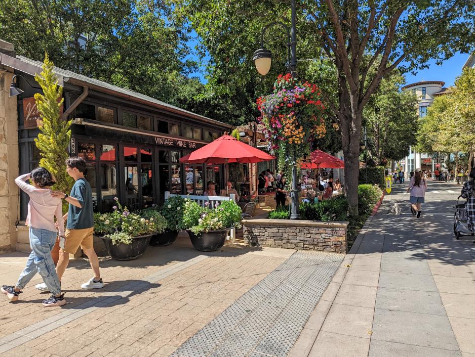 Photo of people walking in front of a smaller restaurant building with people sitting outside with a lot of landscaping on a sunny day