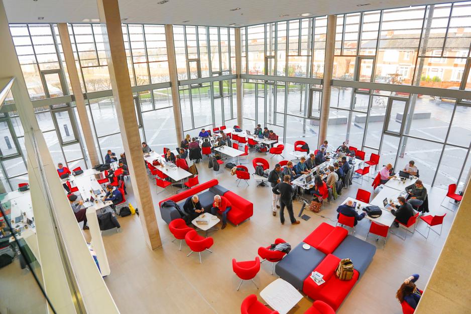 Photo taken from higher up looking down on the lobby of a building filled with red and black tables, chairs, and couches and people sitting and working, talking, or relaxing