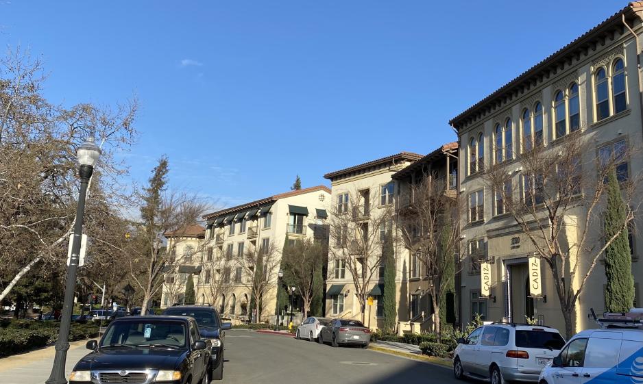 Photo of taller tan residential building with trees in front on a sunny day. There are vehicles parked on either side of the street in front of the building