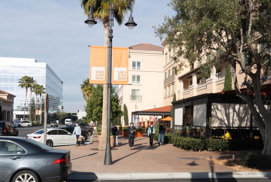 Photo of people walking on a wide sidewalk with outdoor restaurant seating on one side with trees between the sidewalk and street on the other side