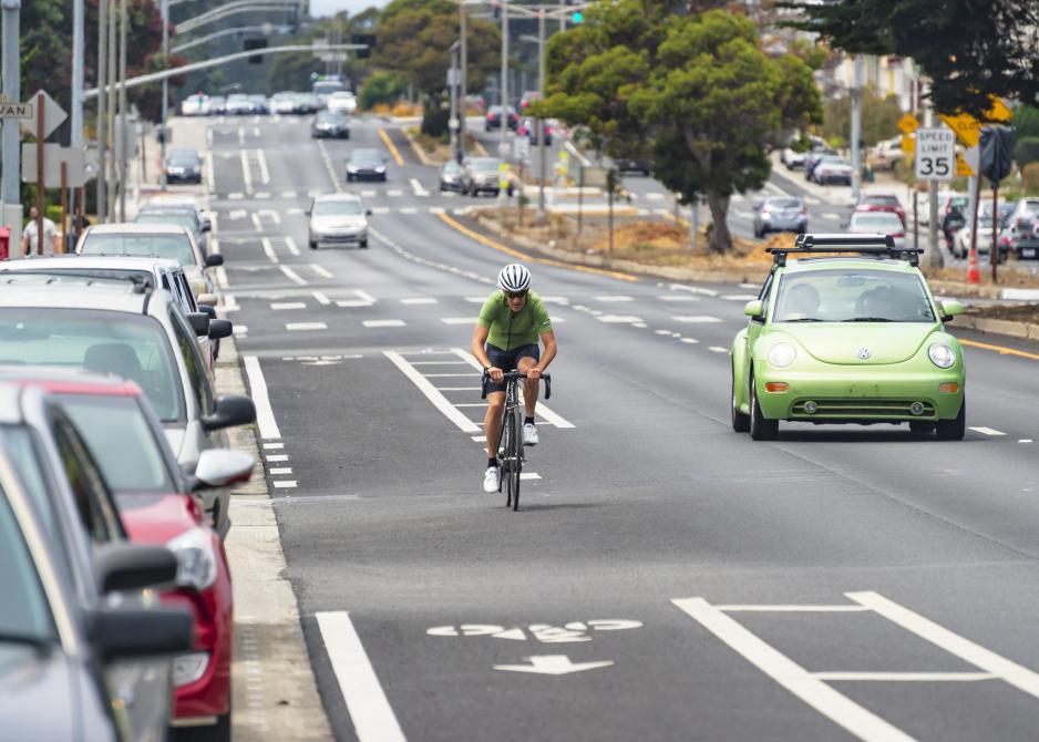 Photo of a multi-lane roadway with cars parked on one side and a bicyclist in the bike lane and a green car in a travel lane