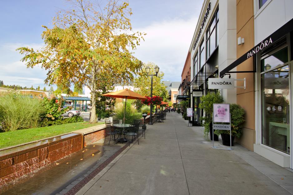 Photo of a sidewalk between colorful shops and raised landscaping and bistro tables 