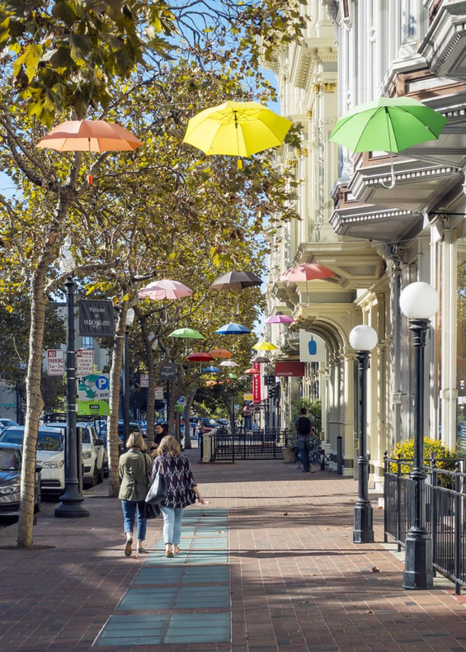 Photo of a sidewalk with people walking under a tree canopy and colorful umbrellas hanging