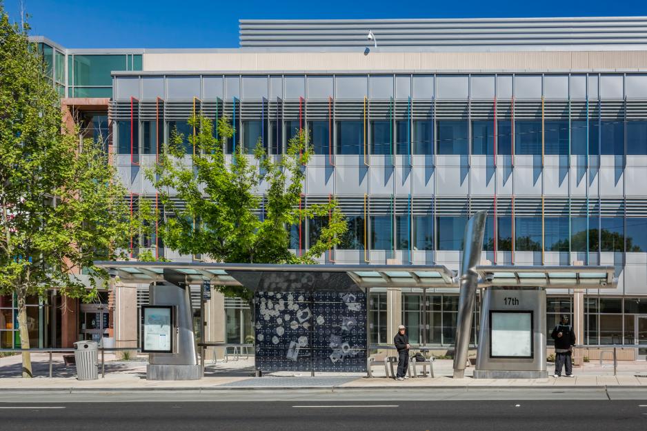 Photo of building with colorful panels partially covering the windows with trees and an artistic bus stop in front