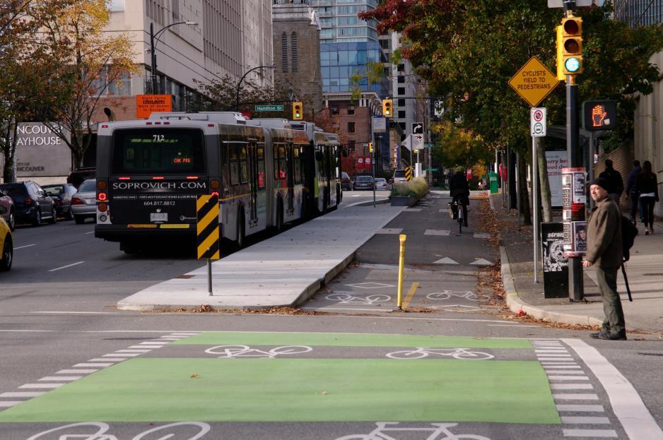 Photo of a city street with a bus stopped at a bus boarding island and a bicyclist riding on a bikeway that wraps behind the island.