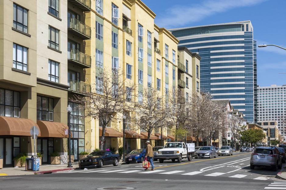 Photo of a city street with street trees, mixed use buildings, a pedestrian crossing the crosswalk.