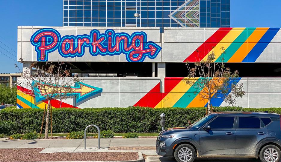 Photo of a two-story parking garage with "parking" written on the side in large, brightly-colored letters with arrows and stripes. There are smaller trees and a parked car in front of the garage