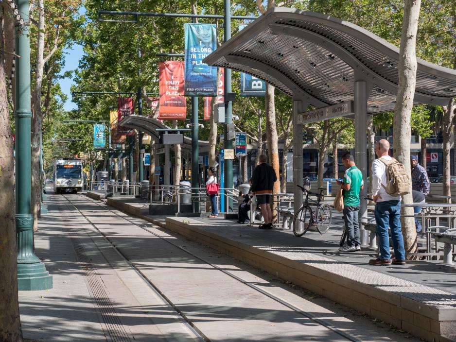 Photo of a light rail station with many people standing on the platforms as a train approaches from the left. There are many trees on both sides of the tracks