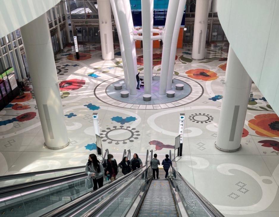 Photo looking down from the top of an escalator onto a level of a train station with people walking and colorful art on the ground